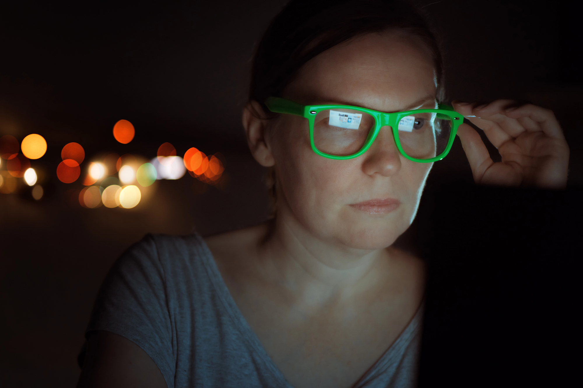 A person with short hair and neon green glasses looks at a screen in a dark setting. The screen light reflects off their glasses. Out-of-focus streetlights in the background create a bokeh effect.