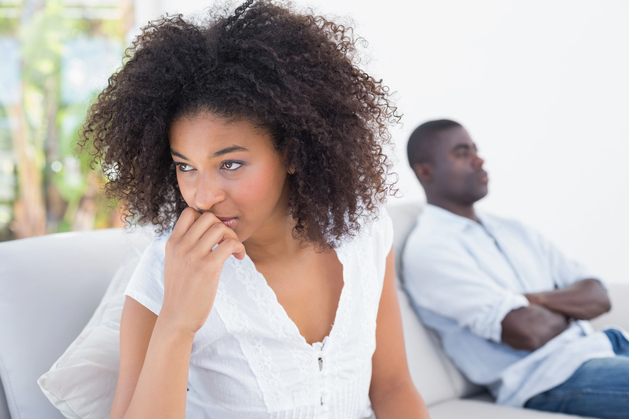 A woman with curly hair looks concerned while sitting on a couch, resting her chin on her hand. A man sits next to her with his arms crossed, looking away. Both appear to be deep in thought.