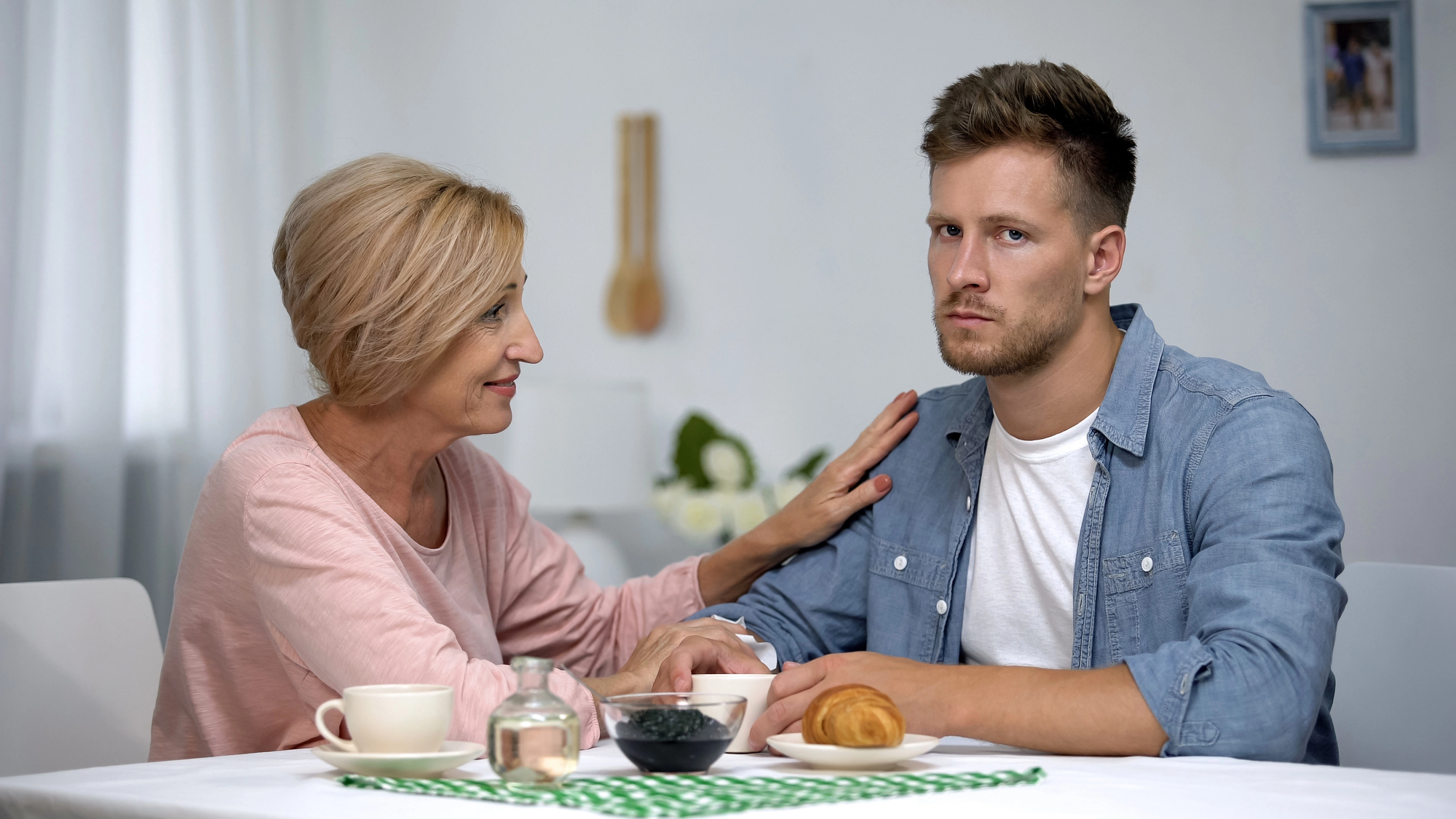 A woman with short blonde hair is sitting at a table, gently touching the shoulder of a man with short brown hair who looks unimpressed. They are in a light-colored room with a meal on the table, including a croissant and drinks.