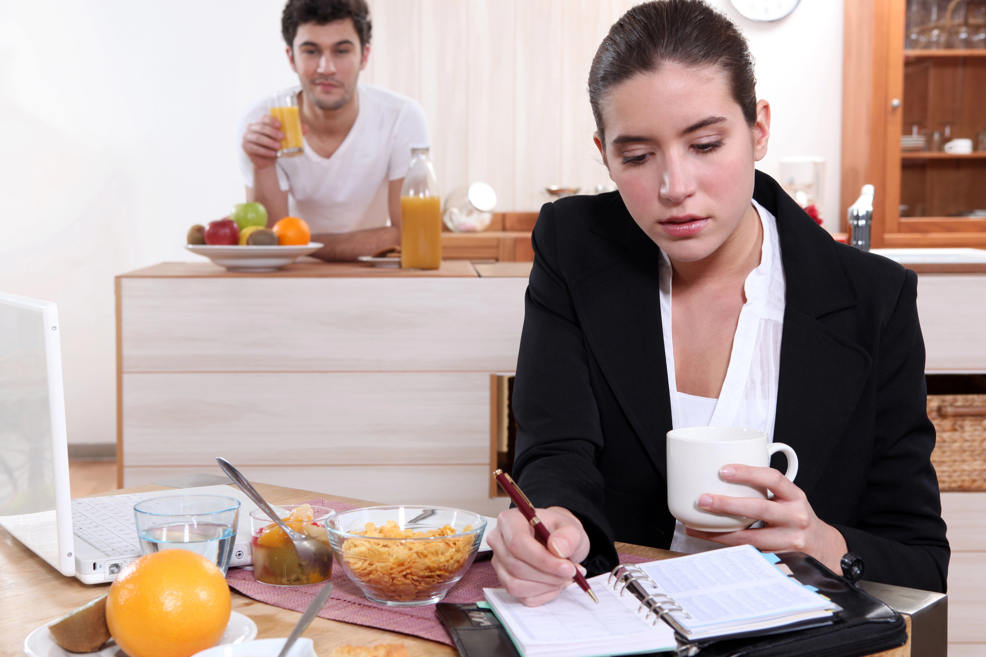 A woman in a black blazer sits at a kitchen table, holding a mug and writing in an organizer. A man in a white shirt stands in the background with a glass of juice. The table is filled with breakfast items like fruits, cereal, and a laptop.