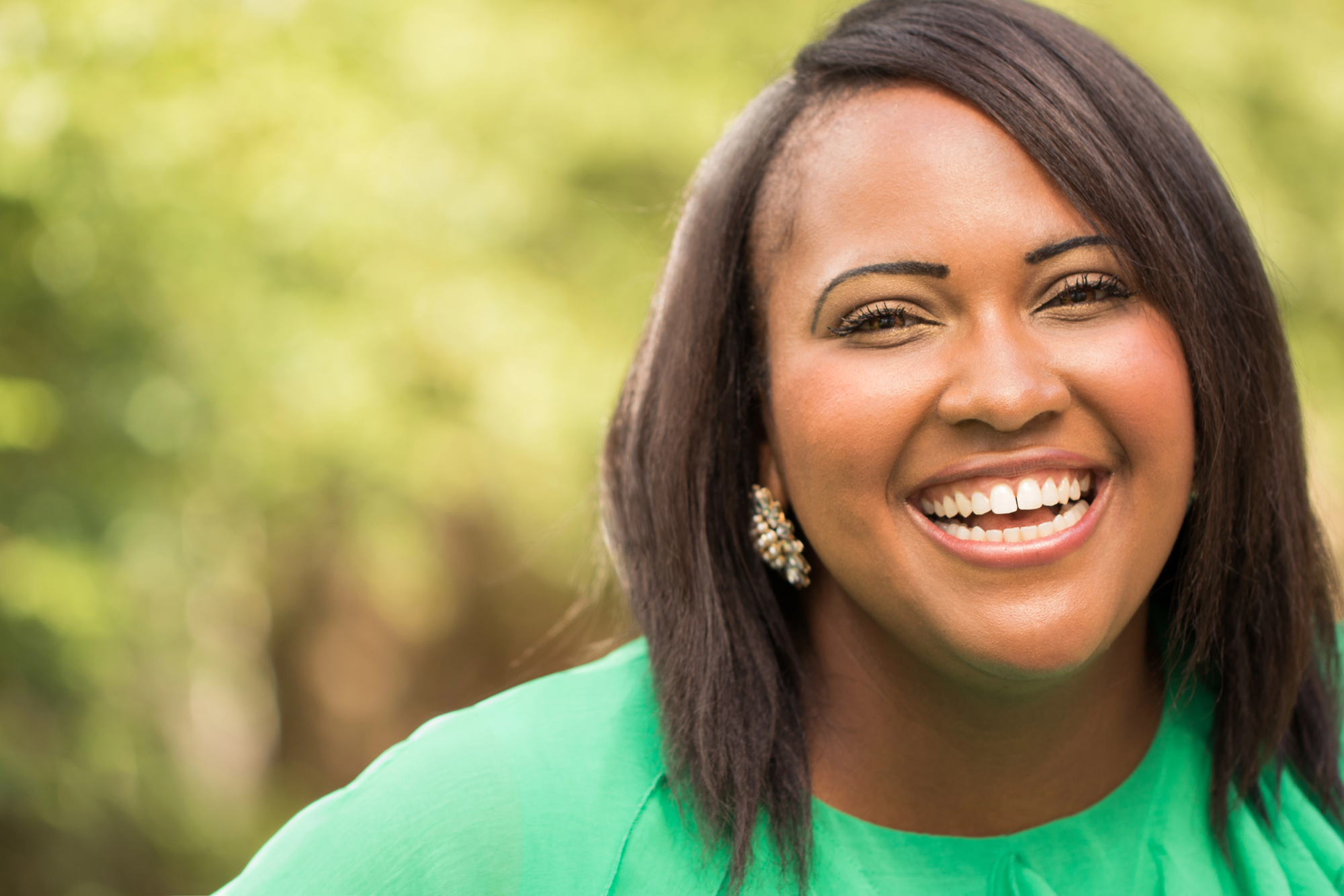 A woman with shoulder-length black hair is smiling brightly. She is wearing a green top and sparkly earrings. The background is a sunny, blurred greenery.