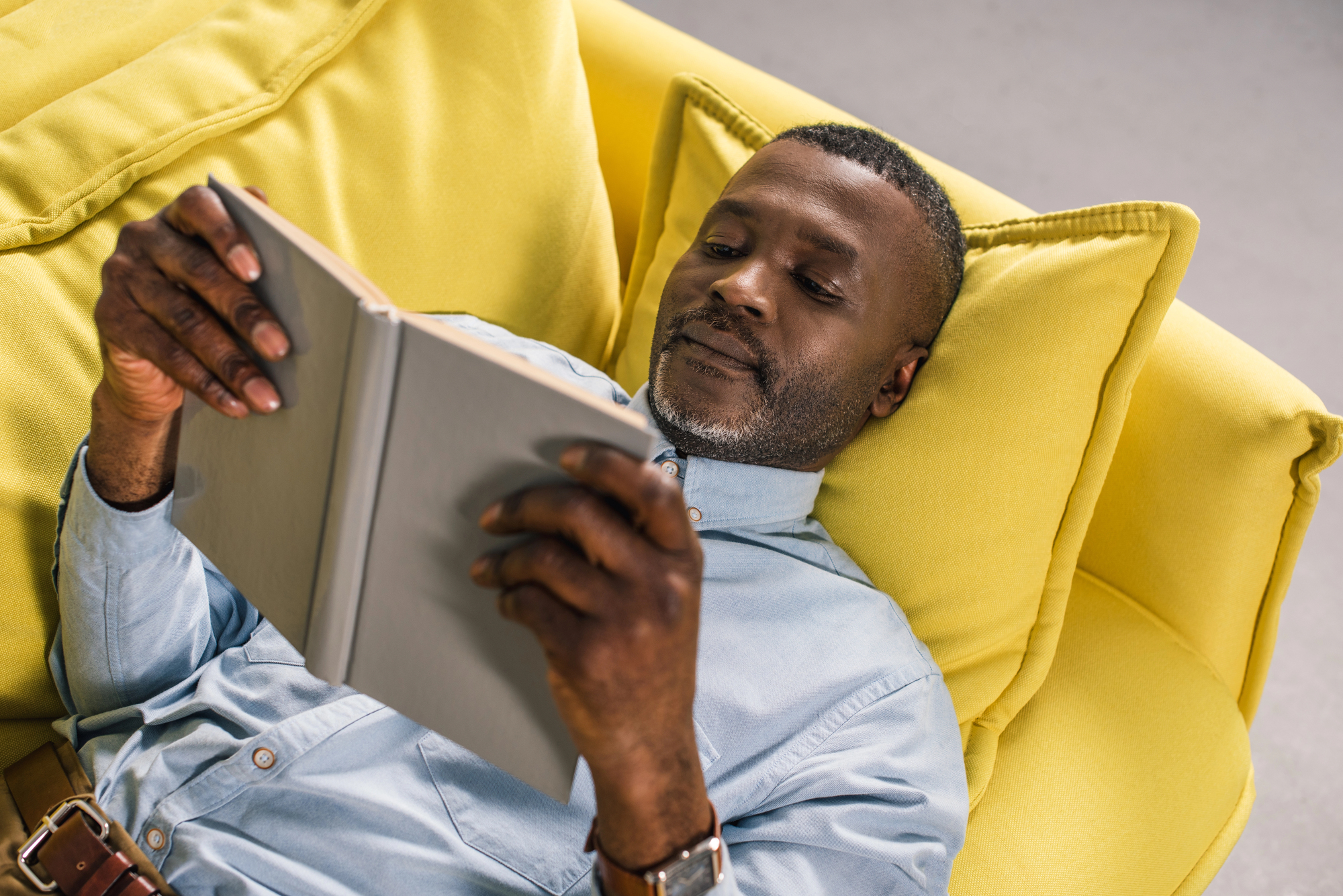 A man is lying comfortably on a yellow couch, reading a book. He is wearing a light blue shirt and appears relaxed, with two yellow cushions supporting him. The background is neutral, emphasizing the cozy indoor setting.