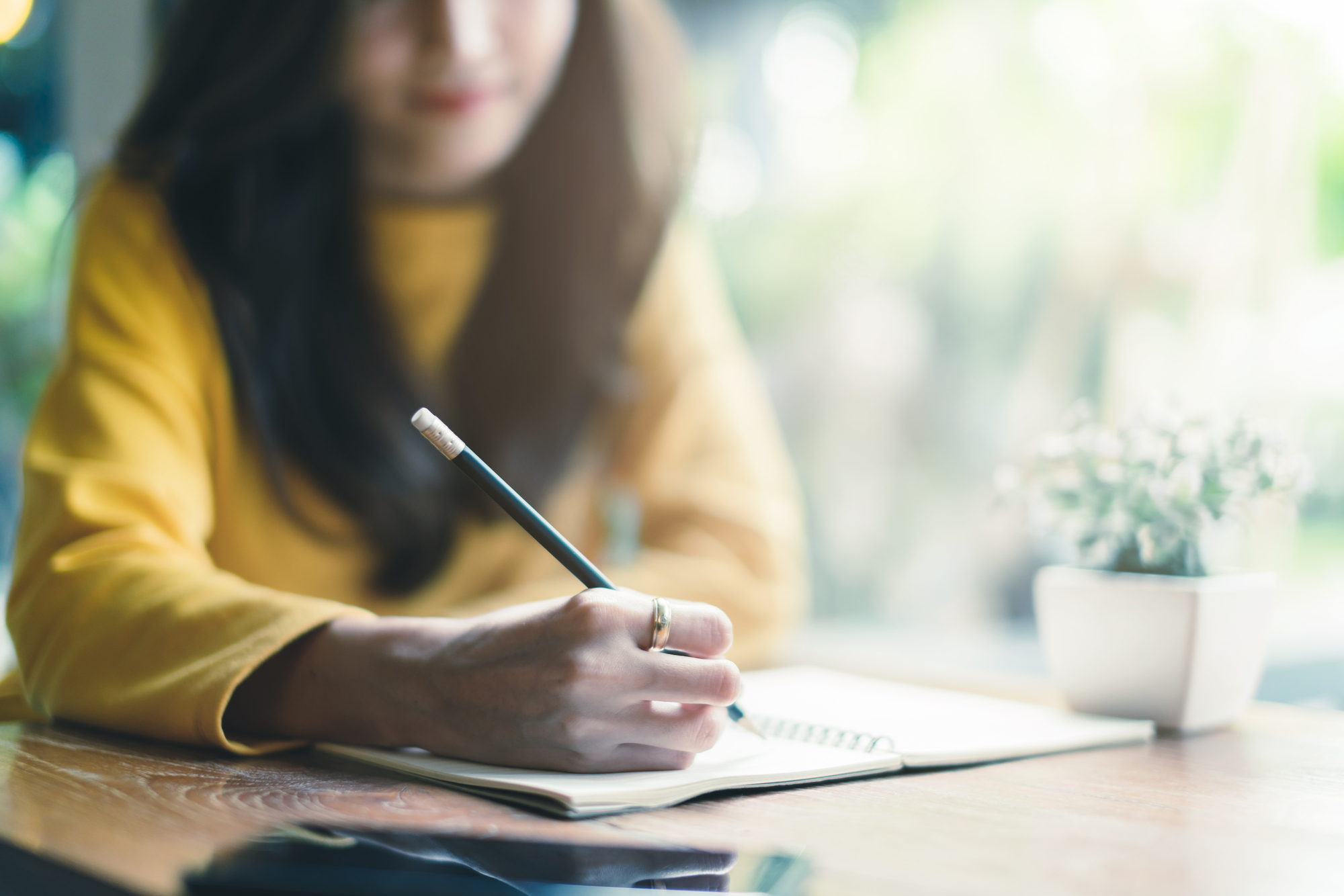 A woman in a yellow sweater writes in a notebook with a pencil at a wooden table. A small potted plant sits nearby, and a phone lies on the table. The background is softly blurred.