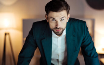 A man with a beard and short hair, wearing a dark green suit jacket and white shirt, leaning forward with a focused expression. Warm interior lighting and blurred background.