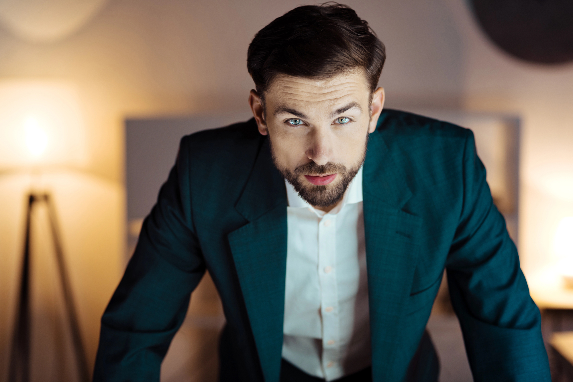 A man with a beard and short hair, wearing a dark green suit jacket and white shirt, leaning forward with a focused expression. Warm interior lighting and blurred background.