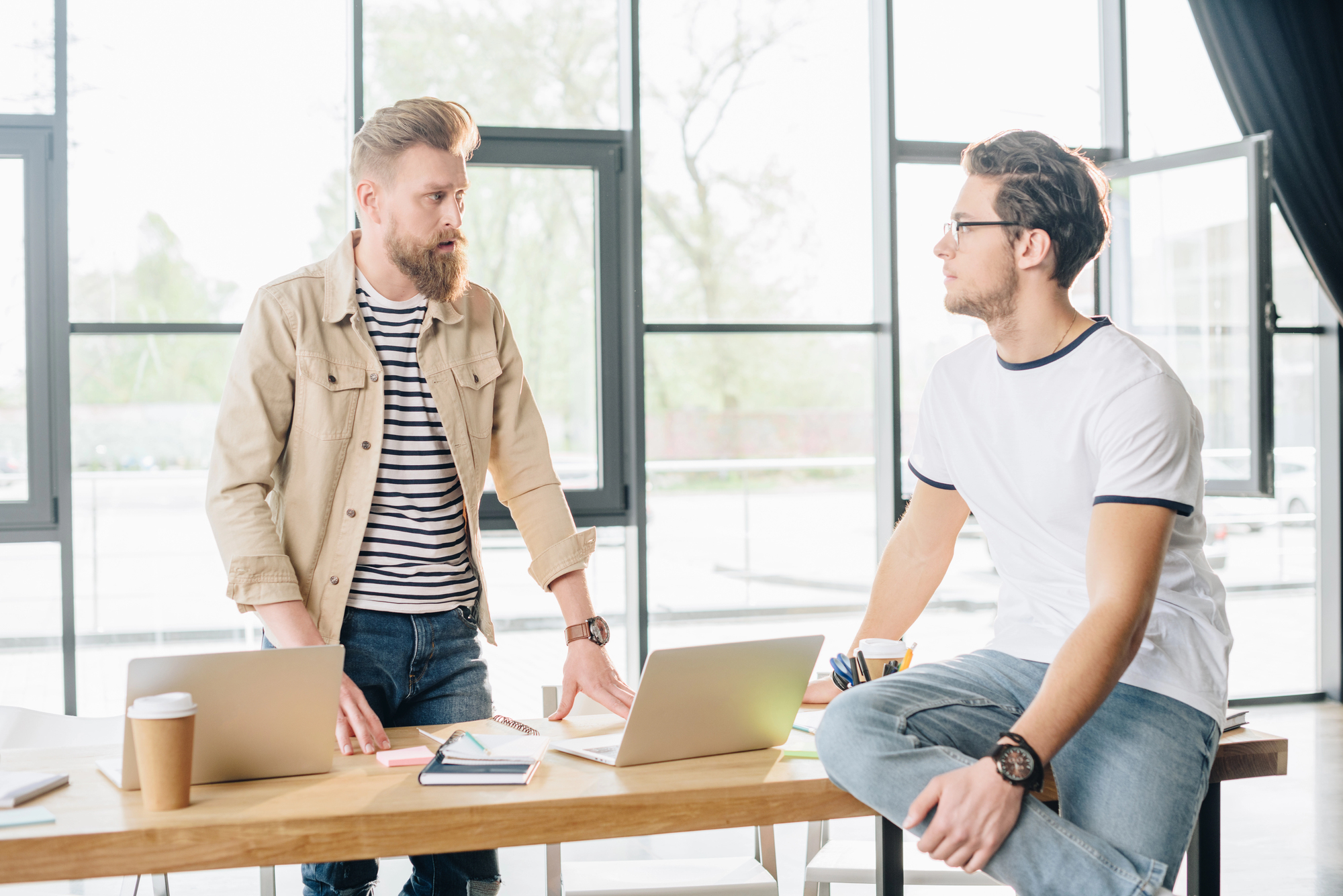 Two men are having a discussion in a bright office. One is standing and wearing a beige jacket, while the other sits on a desk, wearing a white t-shirt. Laptops and a coffee cup are on the wooden table. Large windows are in the background.