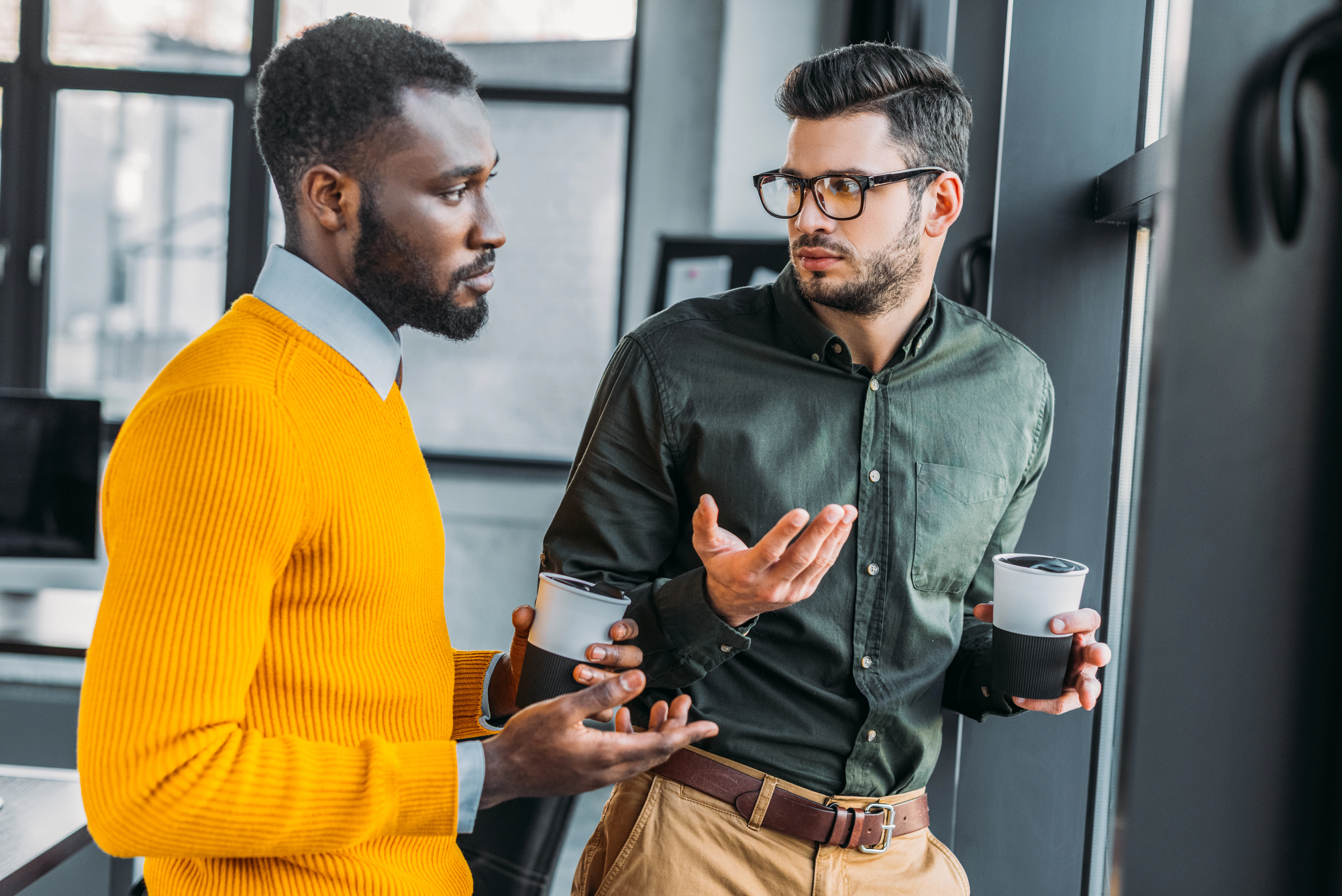 Two men in an office having a conversation while holding coffee cups. One wears a yellow sweater and the other wears a dark green shirt and glasses. They stand near a window, gesturing as they talk.