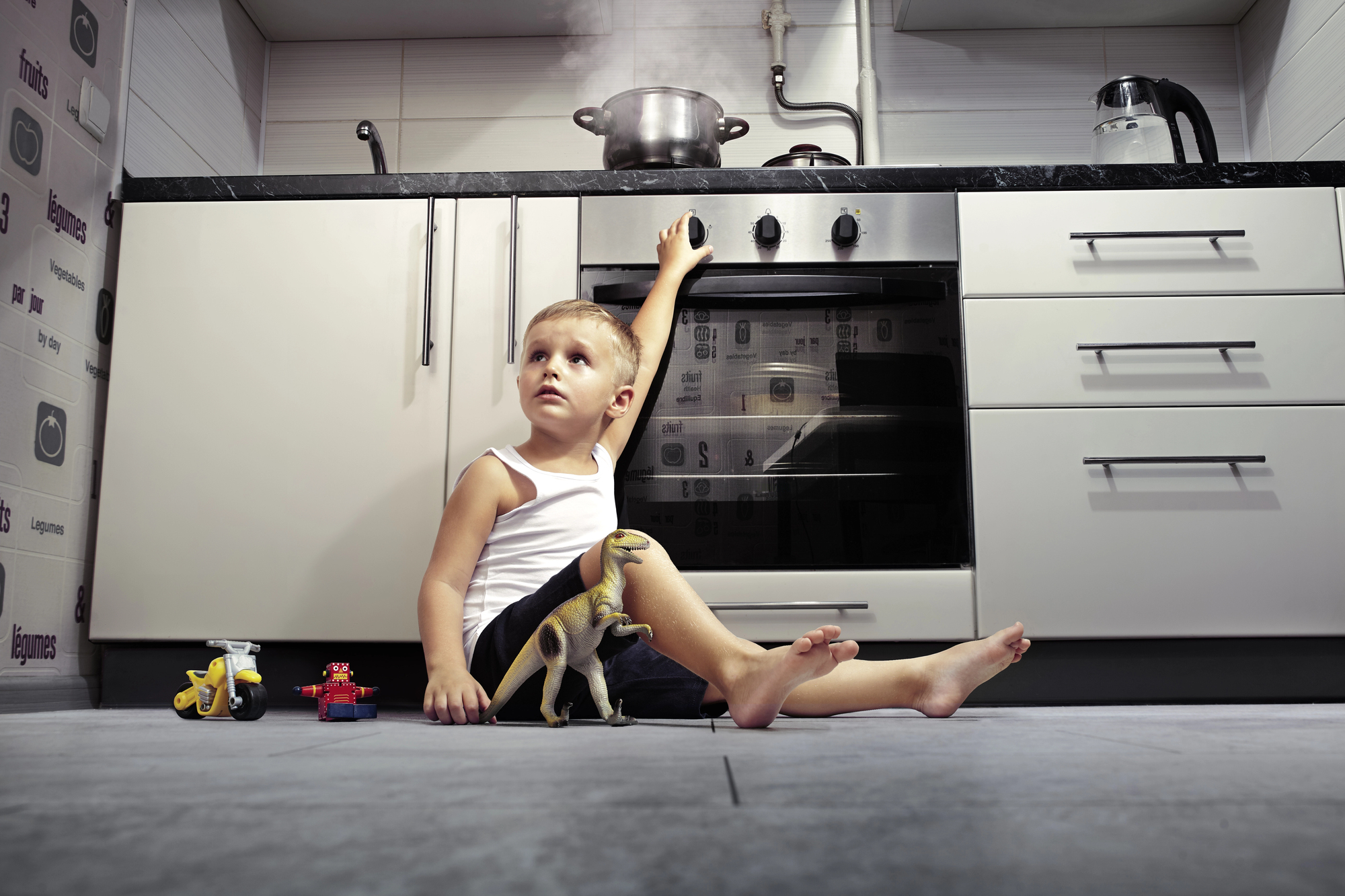 A young boy in a white tank top and shorts is sitting on a kitchen floor, playing with toy trucks and a dinosaur. He is looking up and reaching towards a stove with a pot emitting steam. The kitchen has white cabinets and stainless steel appliances.