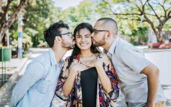 Three friends enjoying a sunny day outdoors. Two men are giving a playful kiss on the cheeks of the smiling woman in the center. They are surrounded by green trees and a bright sky.