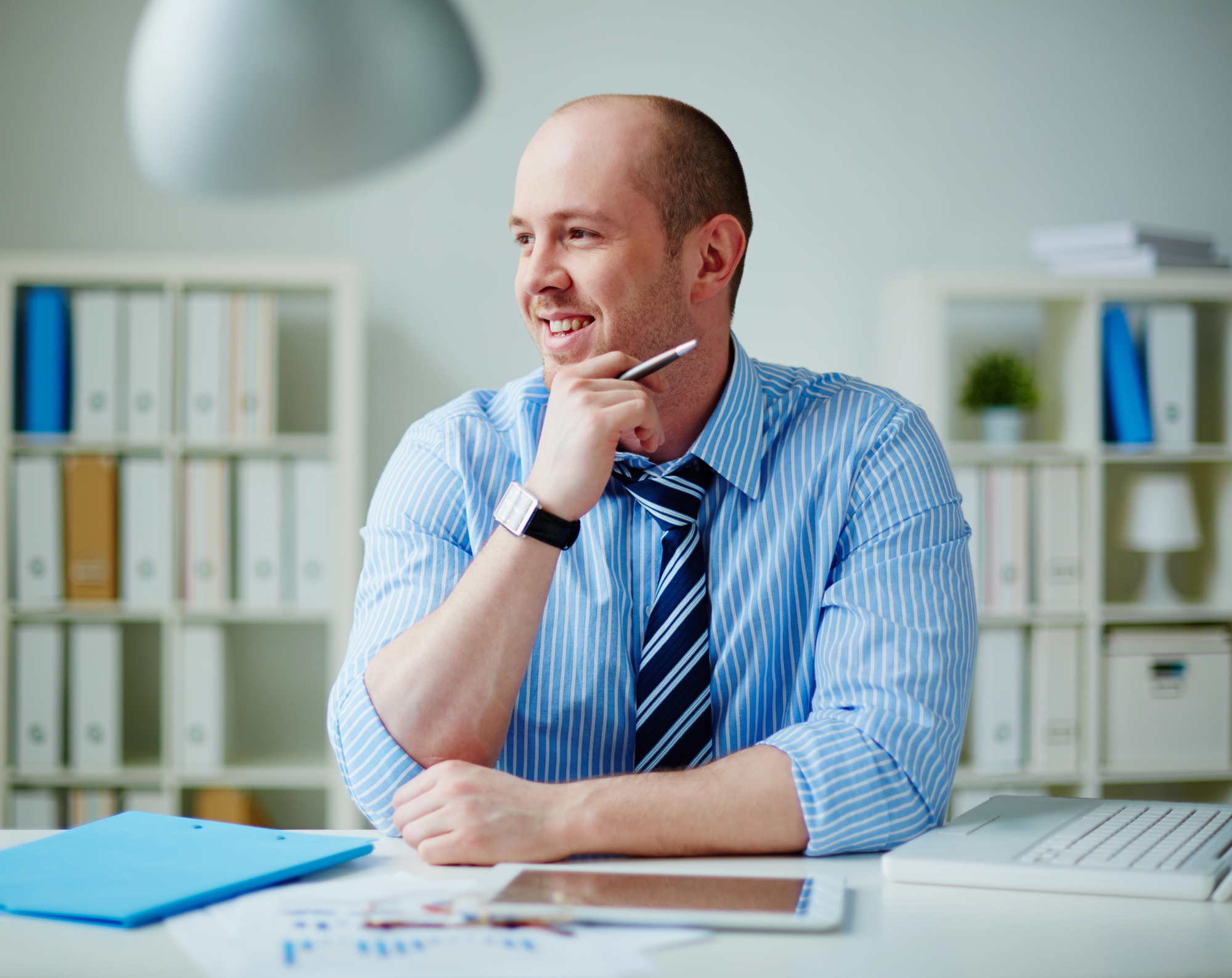 A person seated at a desk, wearing a blue striped shirt and tie, holds a pen and looks to the side with a smile. In the background are shelves with files and decorative items.