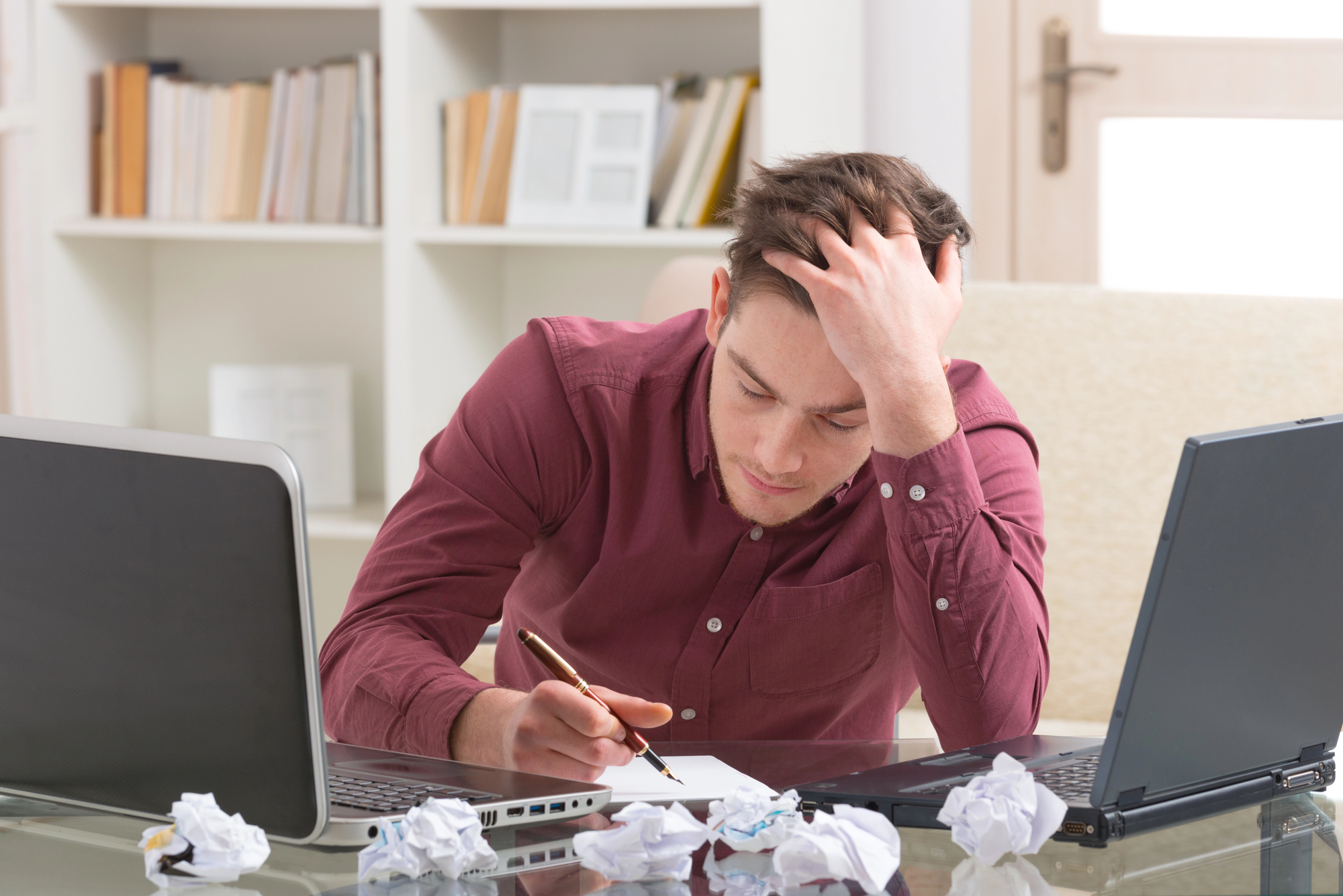 A stressed man sits at a desk with two laptops, surrounded by crumpled papers. He holds his head with one hand while writing on a sheet of paper with the other. Shelves with books and a closed door are visible in the background.