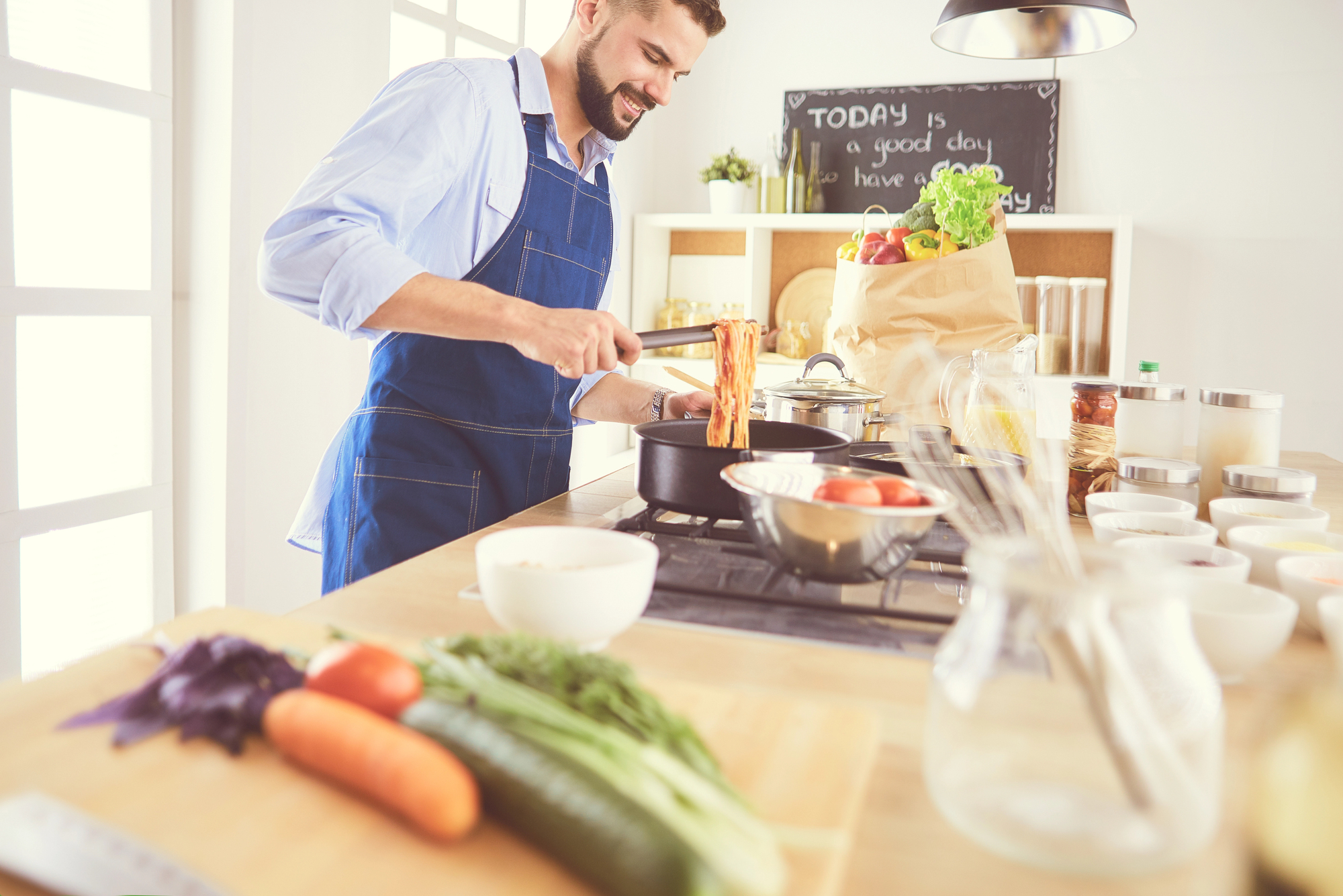 A man in a blue apron is cooking in a bright kitchen. He smiles while stirring food in a pot on the stove. A variety of fresh vegetables, including carrots and zucchini, are on the counter. A chalkboard sign reads, "Today is a good day to have a good day.