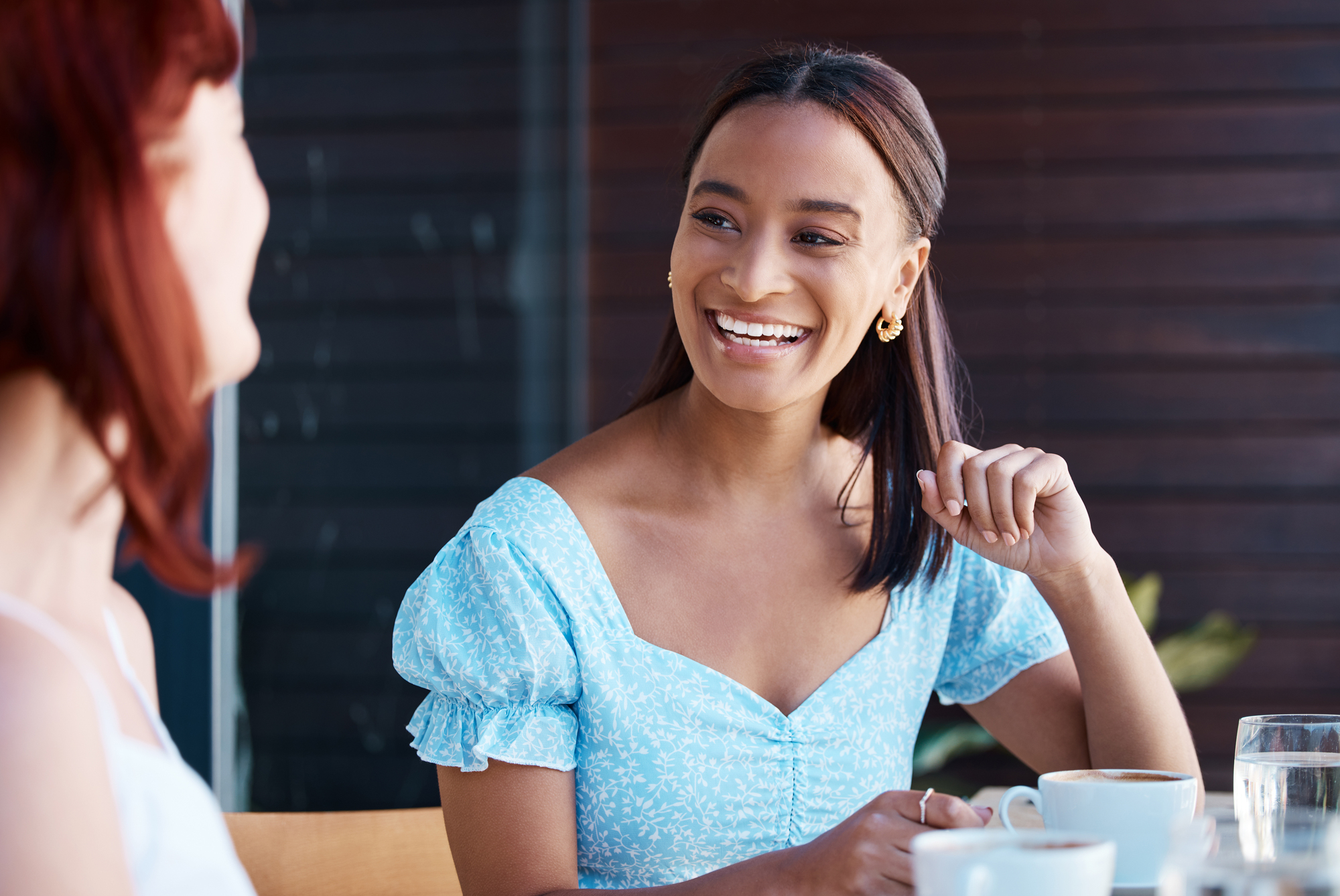 Two women are sitting at a table outdoors, enjoying coffee. One woman with long brown hair is wearing a light blue dress and smiling at the other, who has red hair. The background is a dark wooden panel.