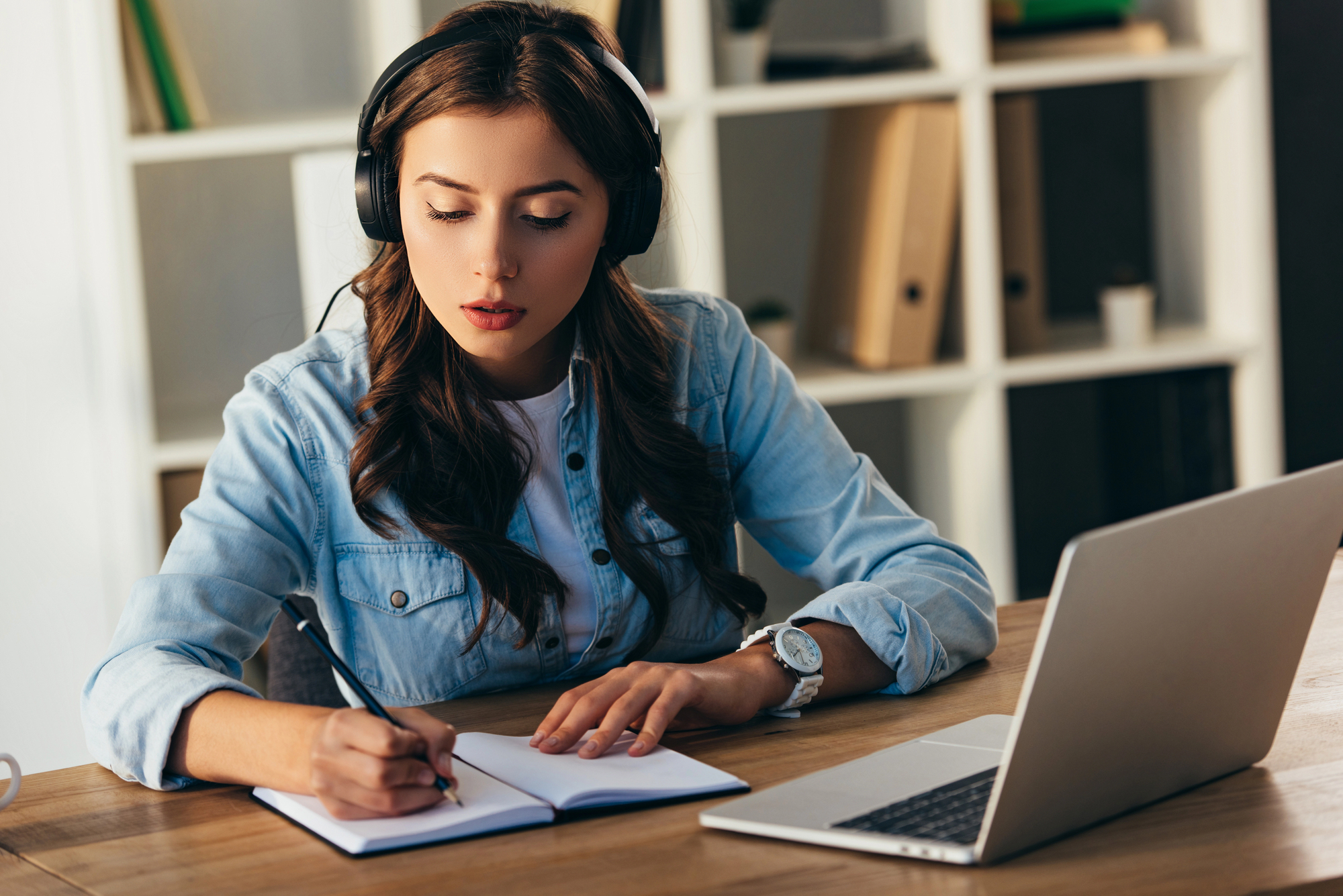 A woman wearing headphones sits at a desk with a laptop. She is writing in a notebook, focused on her work. A shelf with books and binders is in the background.