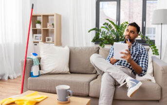 A man sits thoughtfully on a couch, holding a notepad and pen. A mop and cleaning supplies are nearby on the floor. The room is well-lit with a large plant and bookshelf in the background. A coffee cup sits on a table in the foreground.