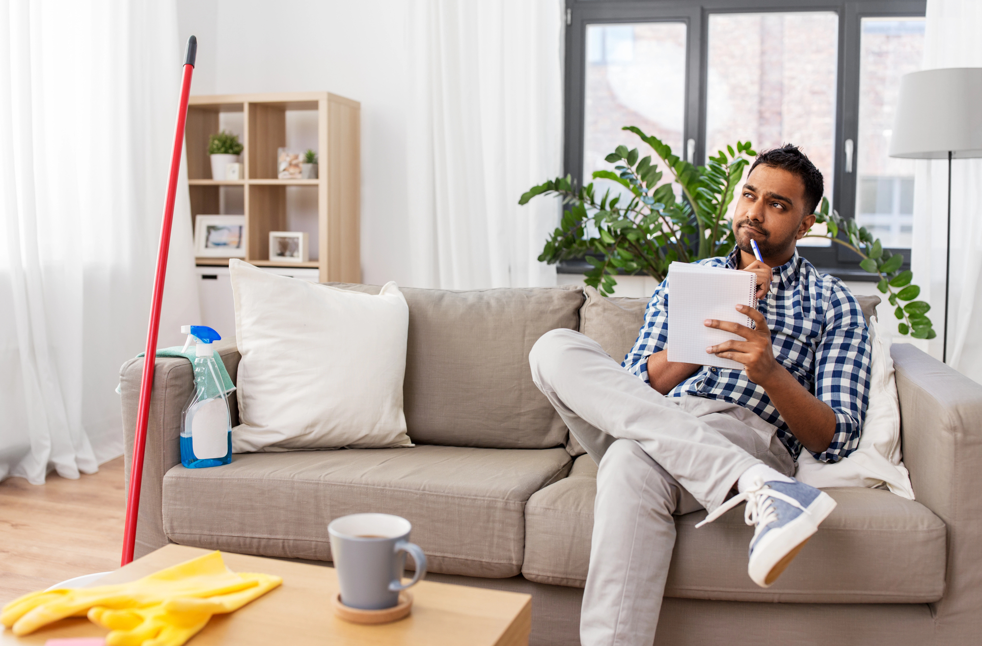 A man sits thoughtfully on a couch, holding a notepad and pen. A mop and cleaning supplies are nearby on the floor. The room is well-lit with a large plant and bookshelf in the background. A coffee cup sits on a table in the foreground.