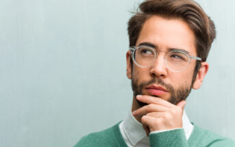 A man with glasses and a beard is wearing a green sweater over a white shirt. He has his hand on his chin and is looking thoughtfully to the side against a plain, light gray background.