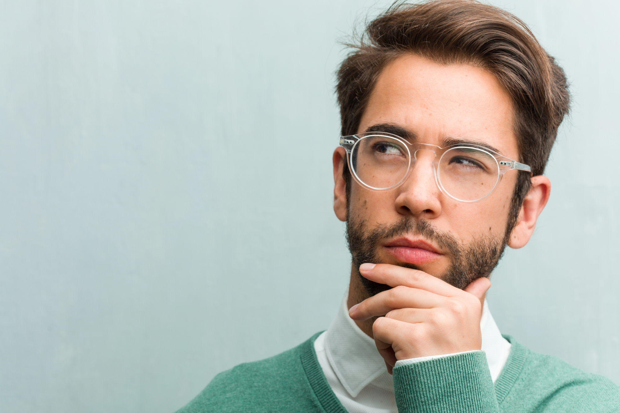 A man with glasses and a beard is wearing a green sweater over a white shirt. He has his hand on his chin and is looking thoughtfully to the side against a plain, light gray background.