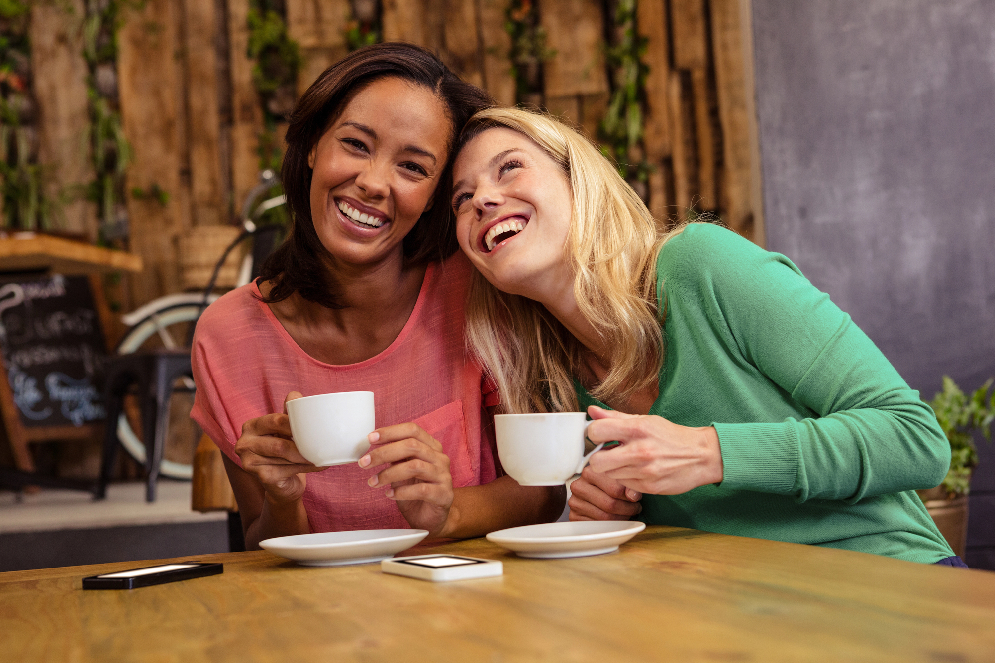 Two women sit together in a cozy cafe, smiling and holding white coffee cups. One woman leans affectionately towards the other. There are smartphones on the wooden table in front of them, and a rustic wall is in the background.