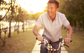 A man in a light gray polo shirt rides a bicycle along a sunlit path lined with trees. He is smiling, and the warm glow of the setting sun filters through the trees, casting a soft light on the scene.