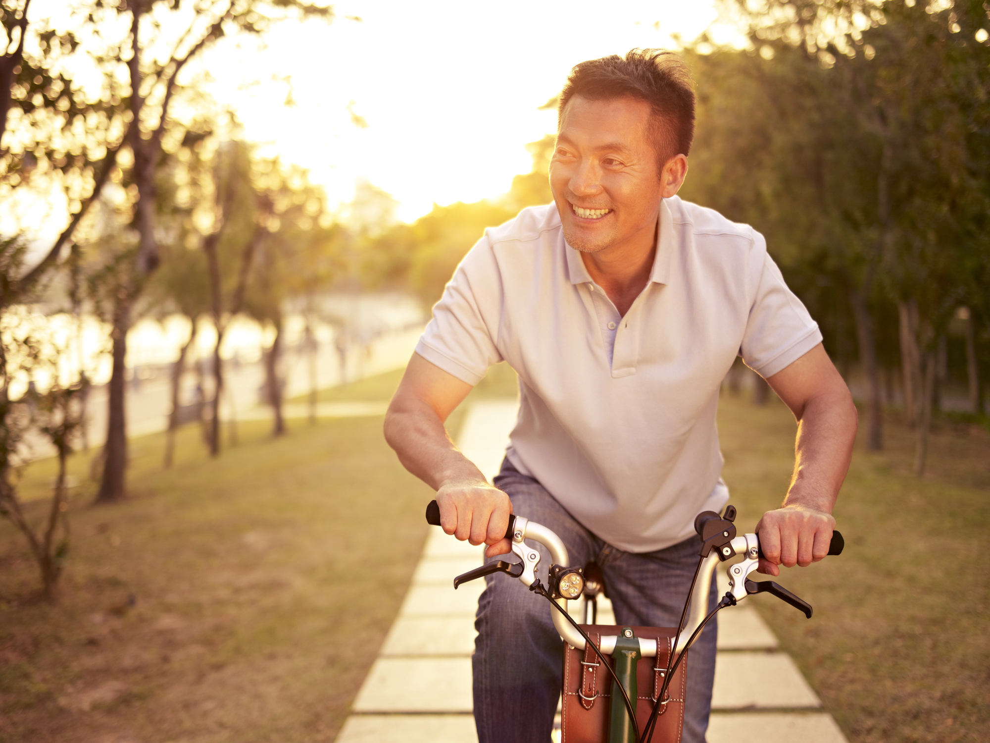 A man in a light gray polo shirt rides a bicycle along a sunlit path lined with trees. He is smiling, and the warm glow of the setting sun filters through the trees, casting a soft light on the scene.