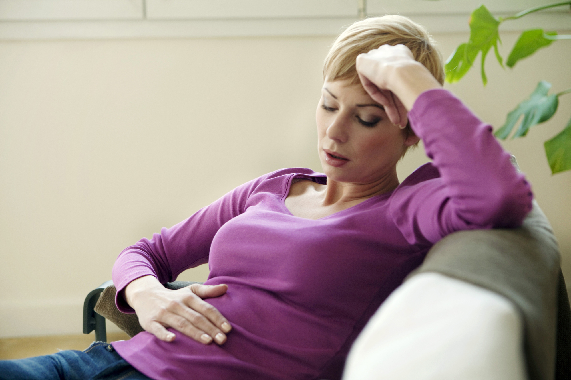 A woman with short blond hair is sitting on a sofa, wearing a purple long-sleeve shirt and blue jeans. She looks unwell, resting her head on her hand and holding her stomach, with a plant visible in the background.