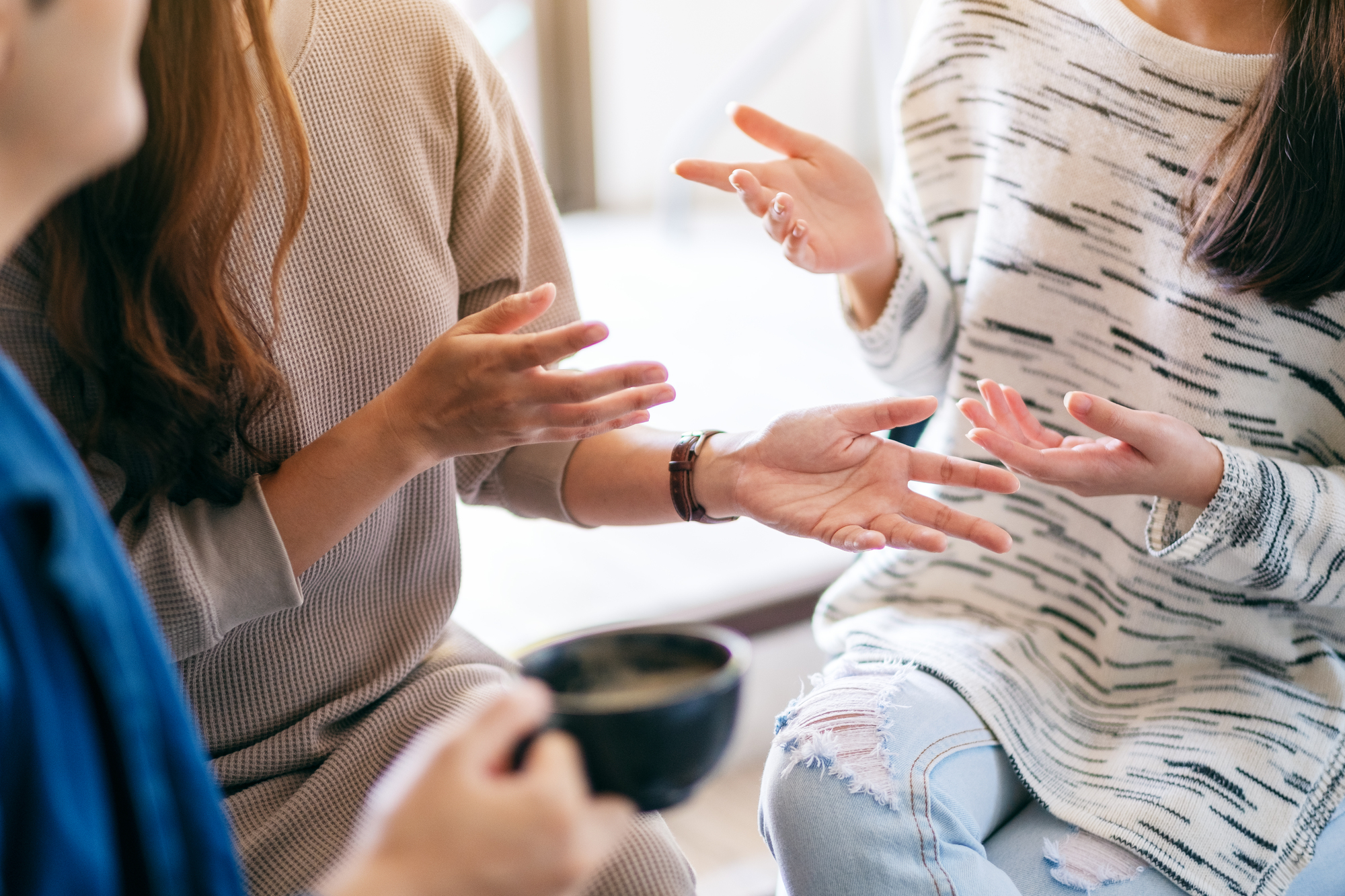 Three people are engaged in a lively conversation. Their hands are gesturing expressively, and one person holds a cup. They are seated closely, suggesting a casual and friendly setting.