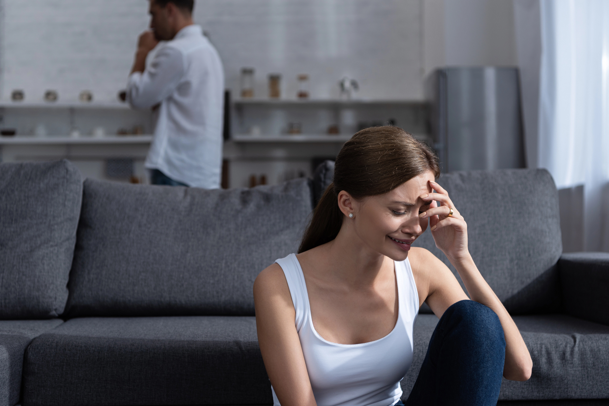 A woman sits on a couch, appearing upset and holding her forehead. In the background, a man stands facing away, looking contemplative. The room is softly lit, with shelves and a window visible.