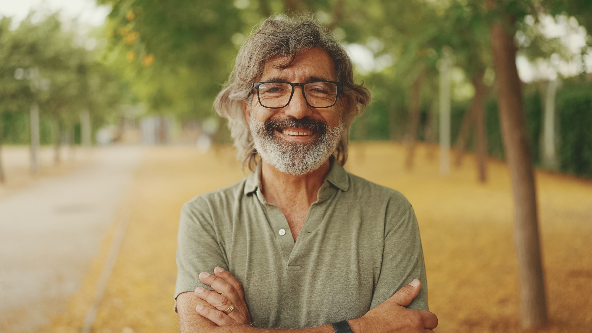 A smiling man with glasses and a gray beard stands with arms crossed in a park. He wears a green polo shirt. The background features trees and a path covered in yellow leaves.