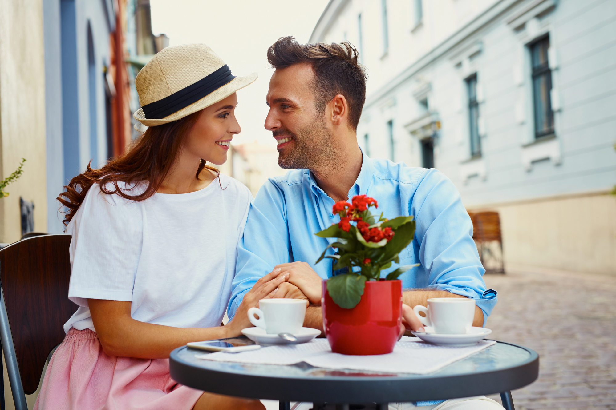 A smiling couple sits at an outdoor café, holding hands across a small table. The woman wears a light outfit and a sunhat; the man is in a light blue shirt. Two cups of coffee are on the table, along with a red potted plant. The background features charming buildings.