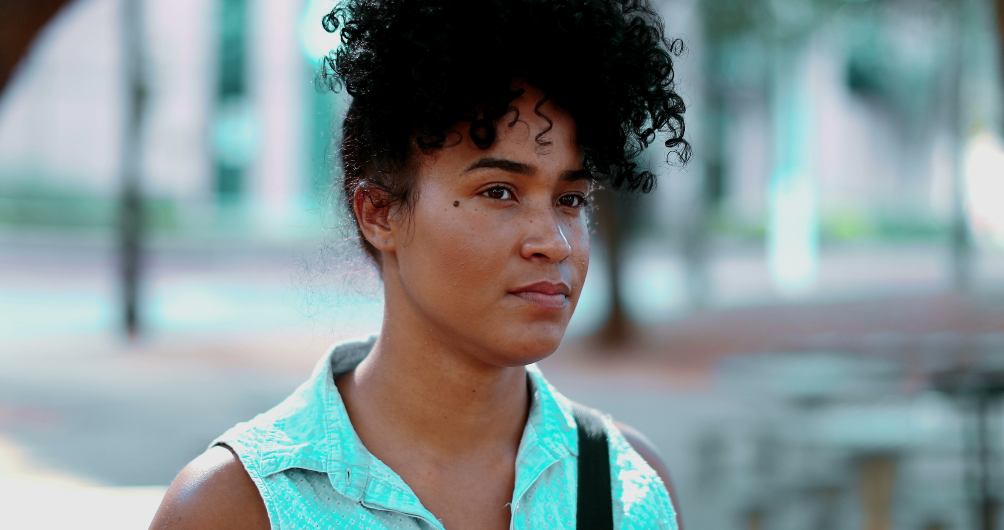 A person with curly black hair looks ahead with a neutral expression. They are wearing a light turquoise sleeveless top. The background is blurred, showing some trees and a paved area.