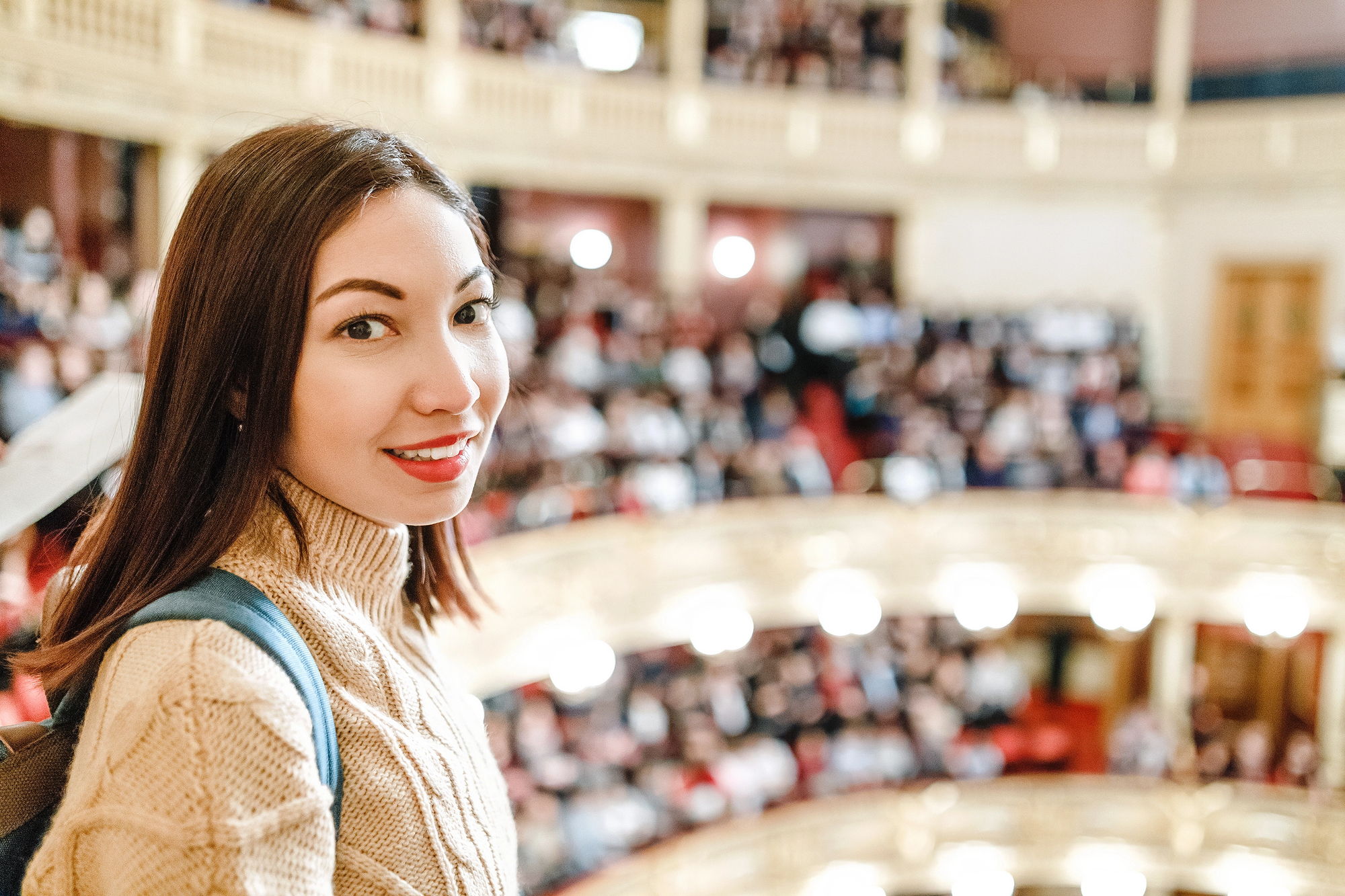 A woman with long hair and a beige sweater stands in an ornate theater. She is smiling, with rows of seats and audience blurred in the background, creating a lively and elegant atmosphere.