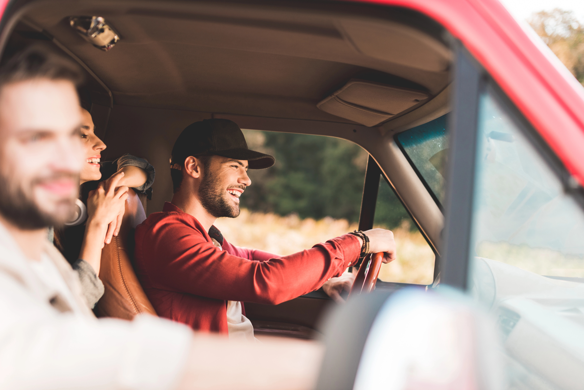 Three people in a red pickup truck are having a good time. The driver, wearing a cap and red shirt, is smiling. Two passengers are seated beside him, one partially visible. They appear to be enjoying a road trip on a sunny day.