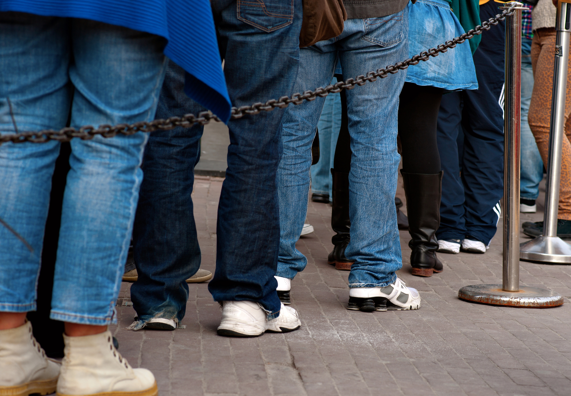 People wearing various types of footwear and jeans stand in line on a cobblestone pavement, separated by metal chains on posts.