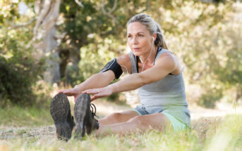 A woman sitting on grassy ground stretches to touch her toes. She is wearing a gray tank top, shorts, and earphones, with an armband on her left arm. Trees and sunlight are visible in the background.