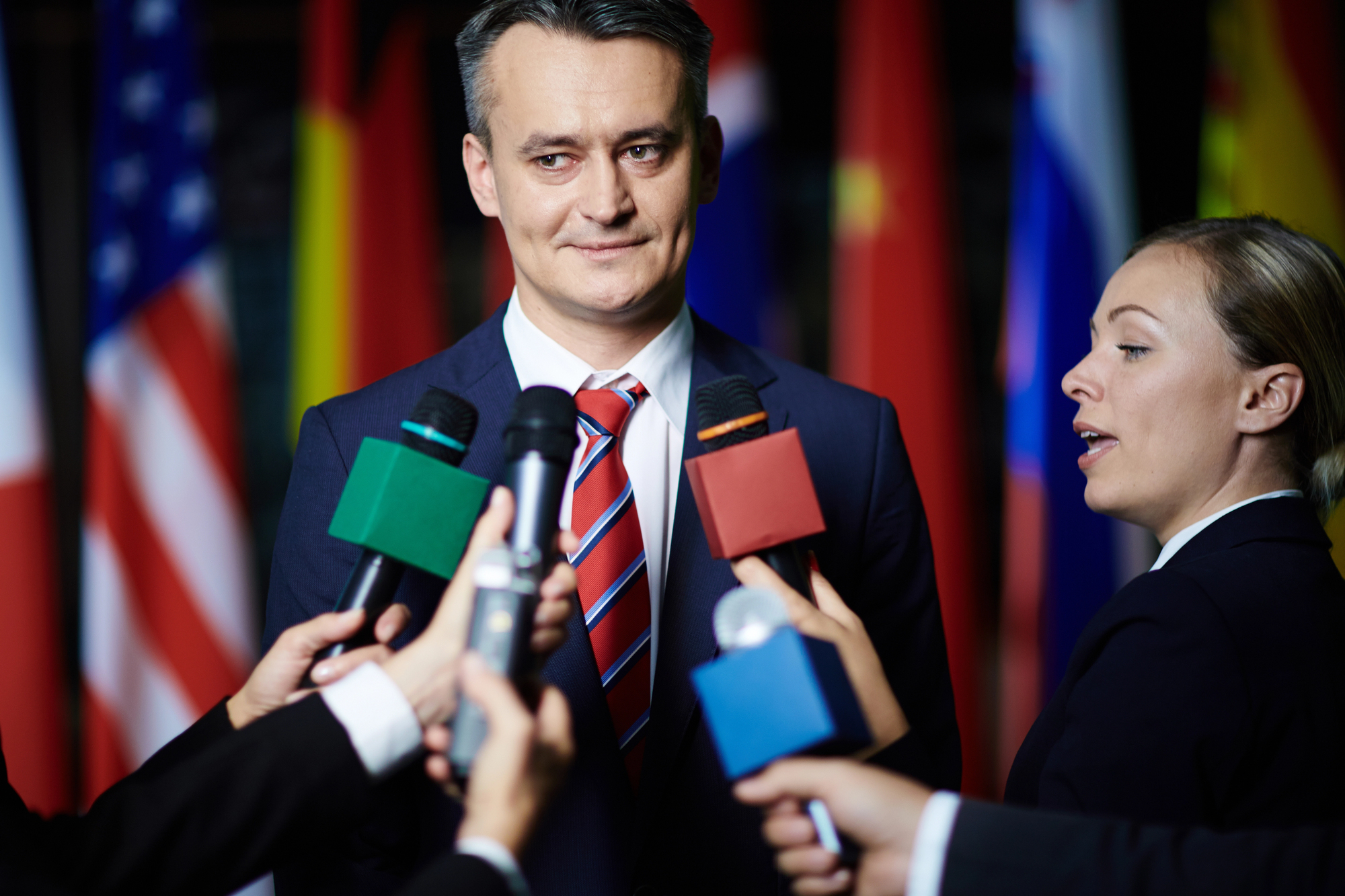 A man in a suit and red striped tie speaks to journalists holding microphones in various colors. Multiple international flags are visible in the background, suggesting a multinational setting.