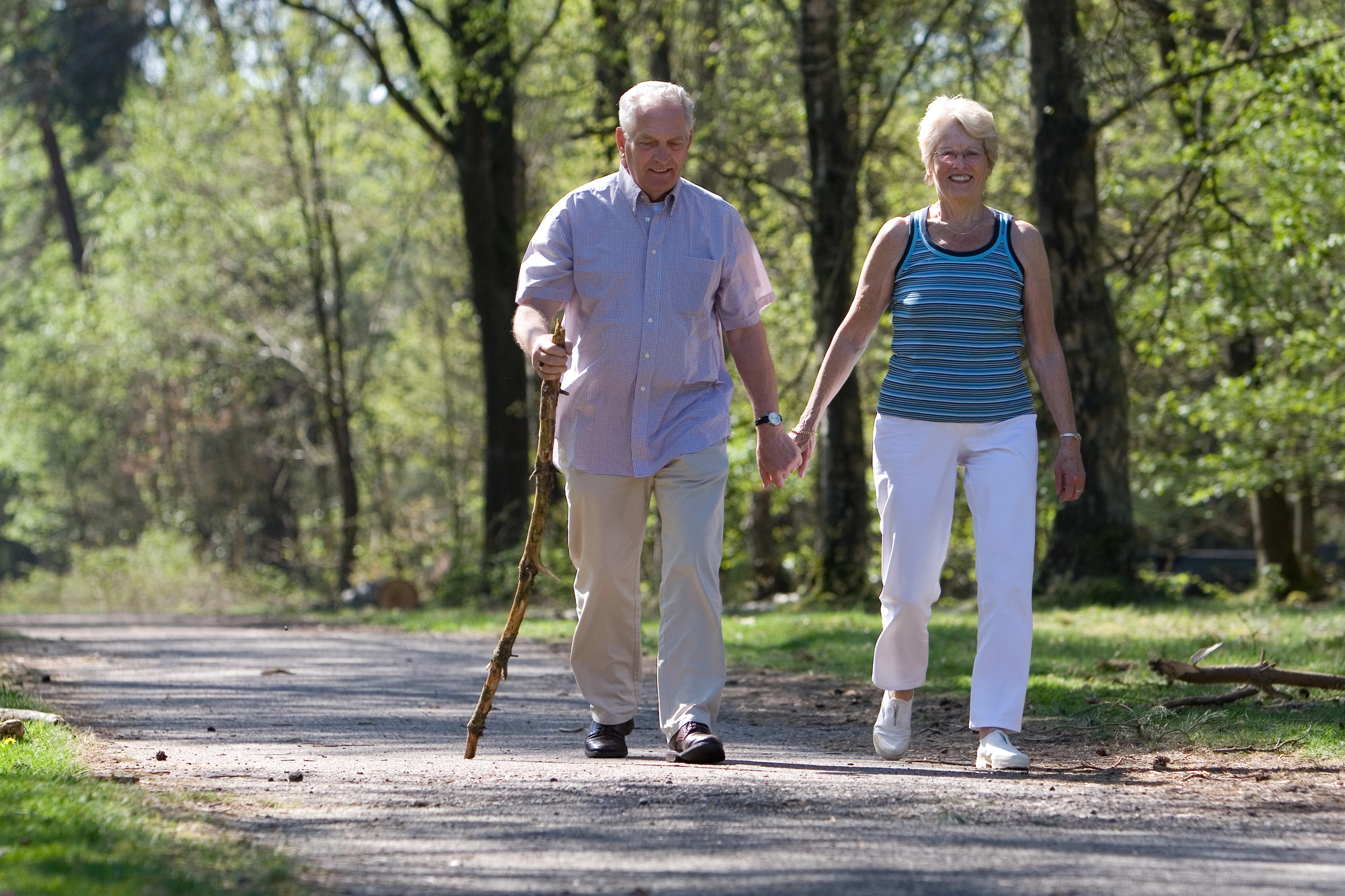 An elderly couple is walking hand in hand on a forest path. The man uses a walking stick and wears a light shirt and trousers, while the woman wears a striped tank top and white pants. Both are smiling, surrounded by lush green trees.