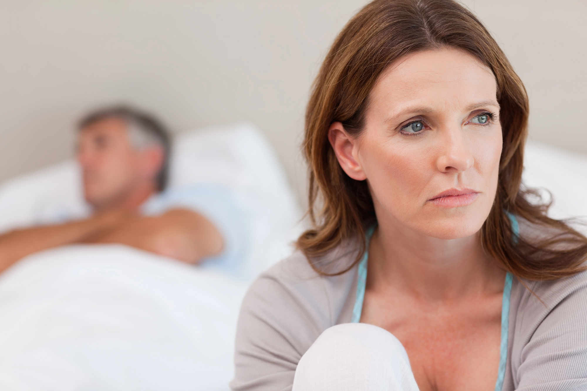 A woman with brown hair looks thoughtfully towards the camera while sitting on a bed. In the background, a man lies in bed, blurred and facing away. The room has a calm, neutral tone, suggesting introspection.