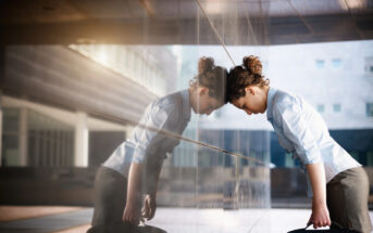 A woman in business attire leans her forehead against a reflective building wall, holding a briefcase. Her expression suggests stress or exhaustion. The urban setting is bright, with modern architecture in the background.