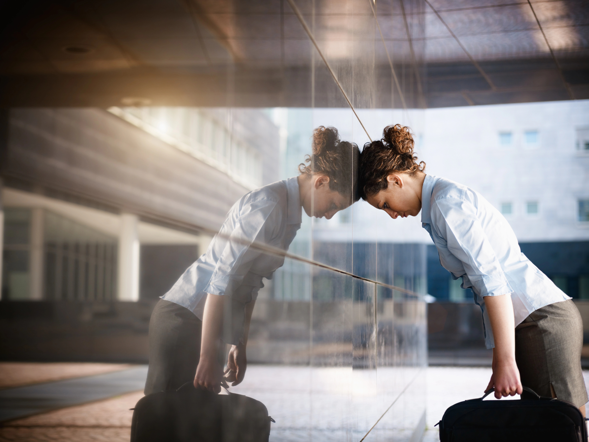 A woman in business attire leans her forehead against a reflective building wall, holding a briefcase. Her expression suggests stress or exhaustion. The urban setting is bright, with modern architecture in the background.