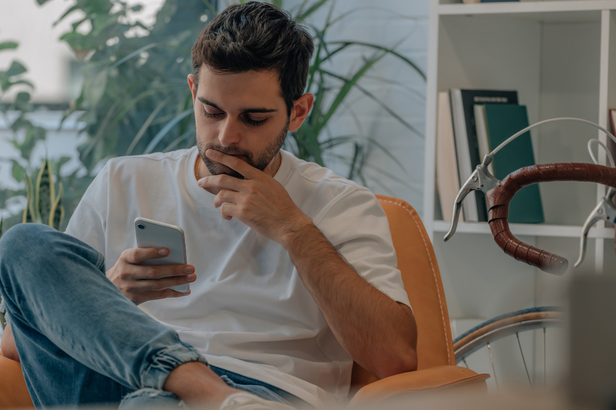 A man sitting on a chair, wearing a white t-shirt and jeans, looking intently at his smartphone with a thoughtful expression. In the background, there is a bicycle and books on a shelf with green plants nearby.