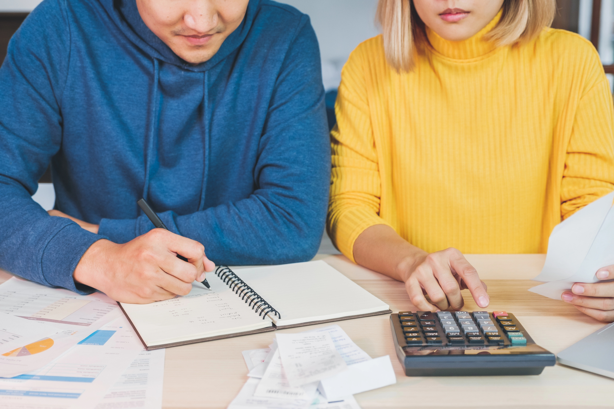 A man in a blue hoodie writes in a notebook while a woman in a yellow sweater uses a calculator. They are surrounded by documents and receipts on a wooden table, focusing on budgeting or financial planning.