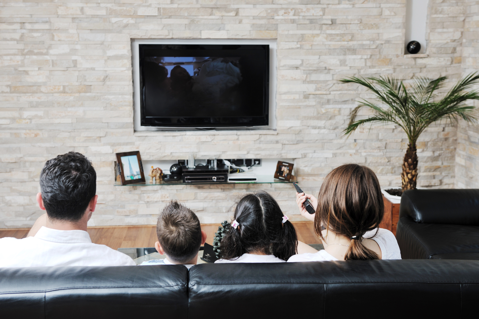 A family of four sits on a black sofa in a living room, watching a large TV mounted on a stone-textured wall. There are decorative items and framed photos on a shelf below the TV, with a potted plant nearby.