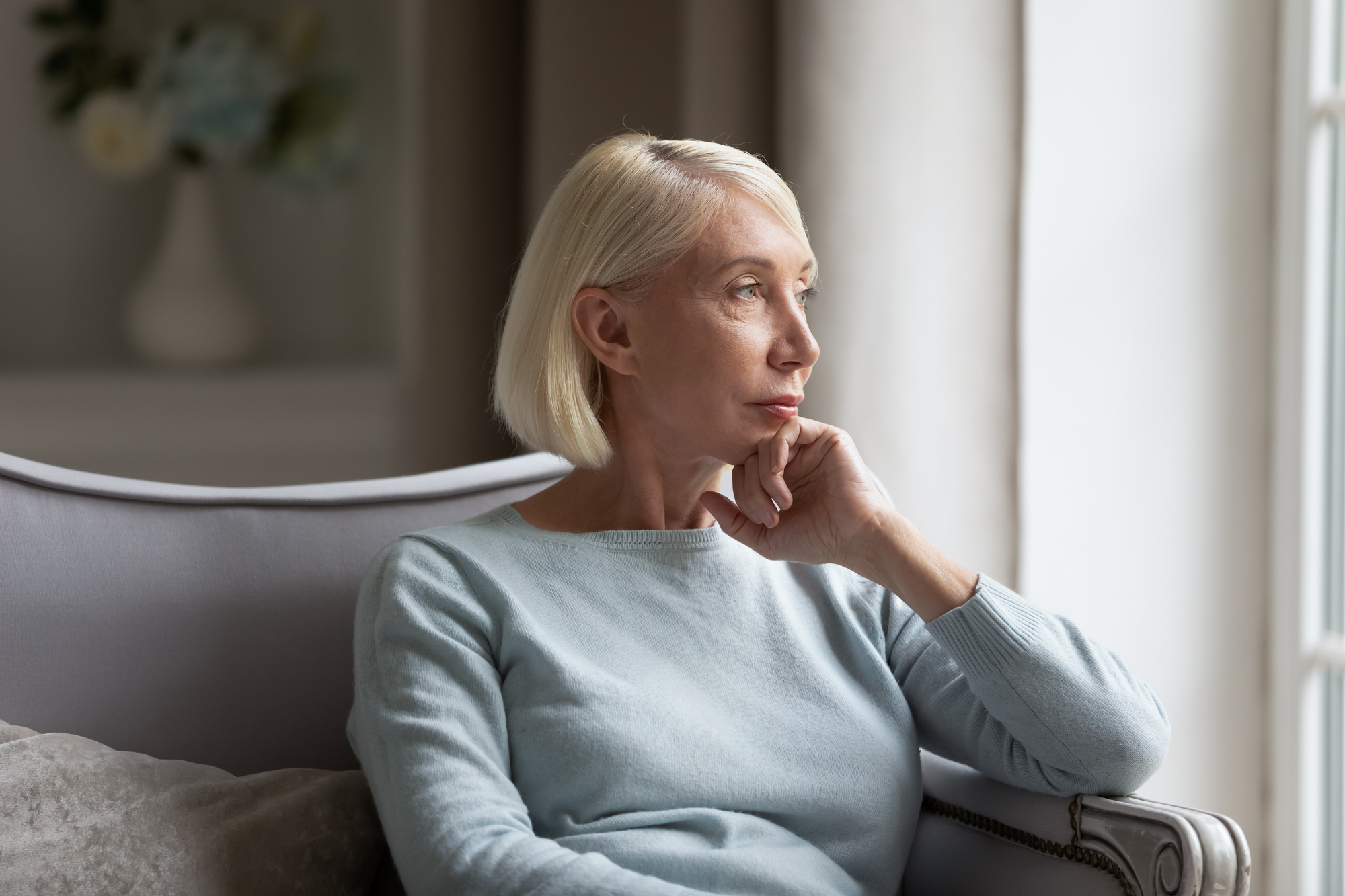 An older woman with short blond hair is sitting on a couch, resting her chin on her hand, and gazing thoughtfully out of a window. She is wearing a light blue sweater and appears contemplative. A blurred vase with flowers is in the background.