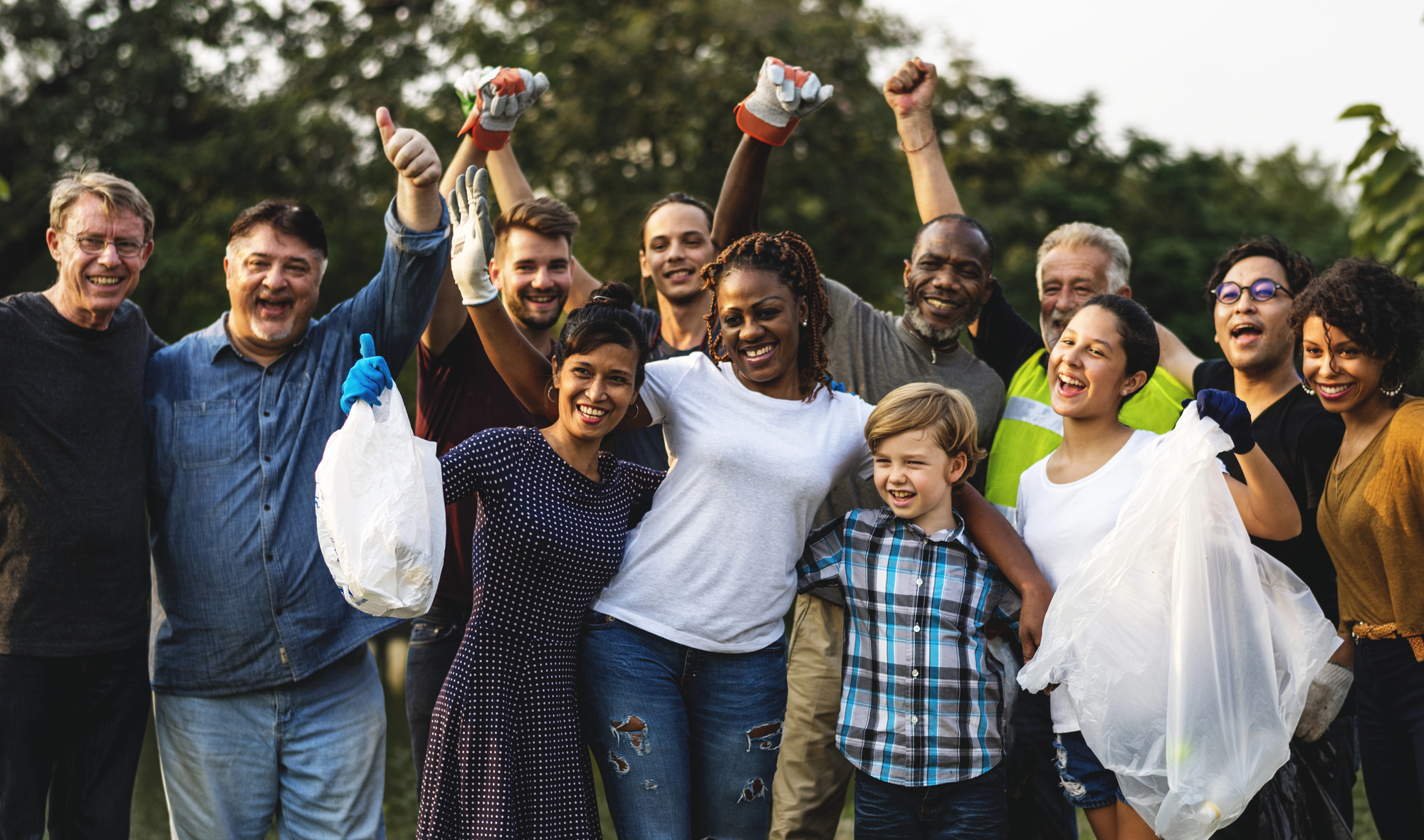 A diverse group of people smiling and celebrating, holding trash bags and wearing gloves, indicating a successful community cleanup. They stand outdoors with trees in the background.