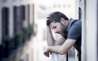 A man leans on a balcony railing, looking thoughtful and resting his head on his hand. He gazes downward, wearing a gray shirt, with an urban building scene in the blurred background.