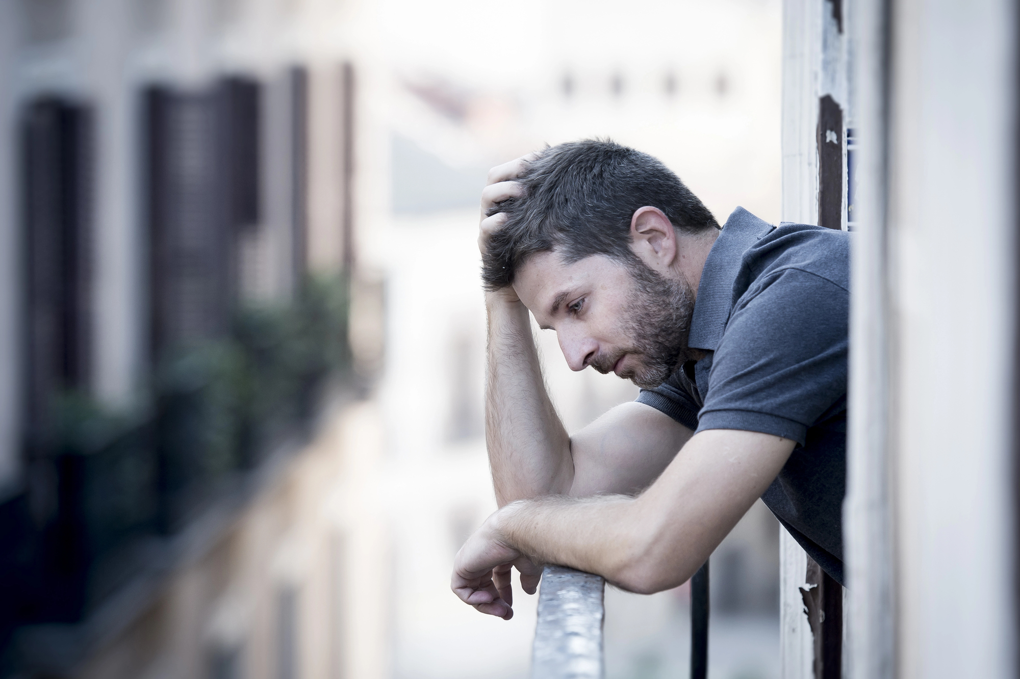 A man leans on a balcony railing, looking thoughtful and resting his head on his hand. He gazes downward, wearing a gray shirt, with an urban building scene in the blurred background.