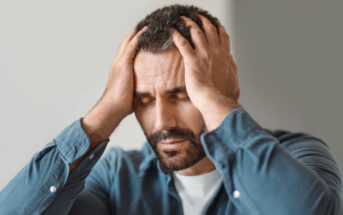 A man with a beard and closed eyes holds his head in his hands, appearing stressed or in pain. He is wearing a blue shirt and is positioned in front of a neutral background.