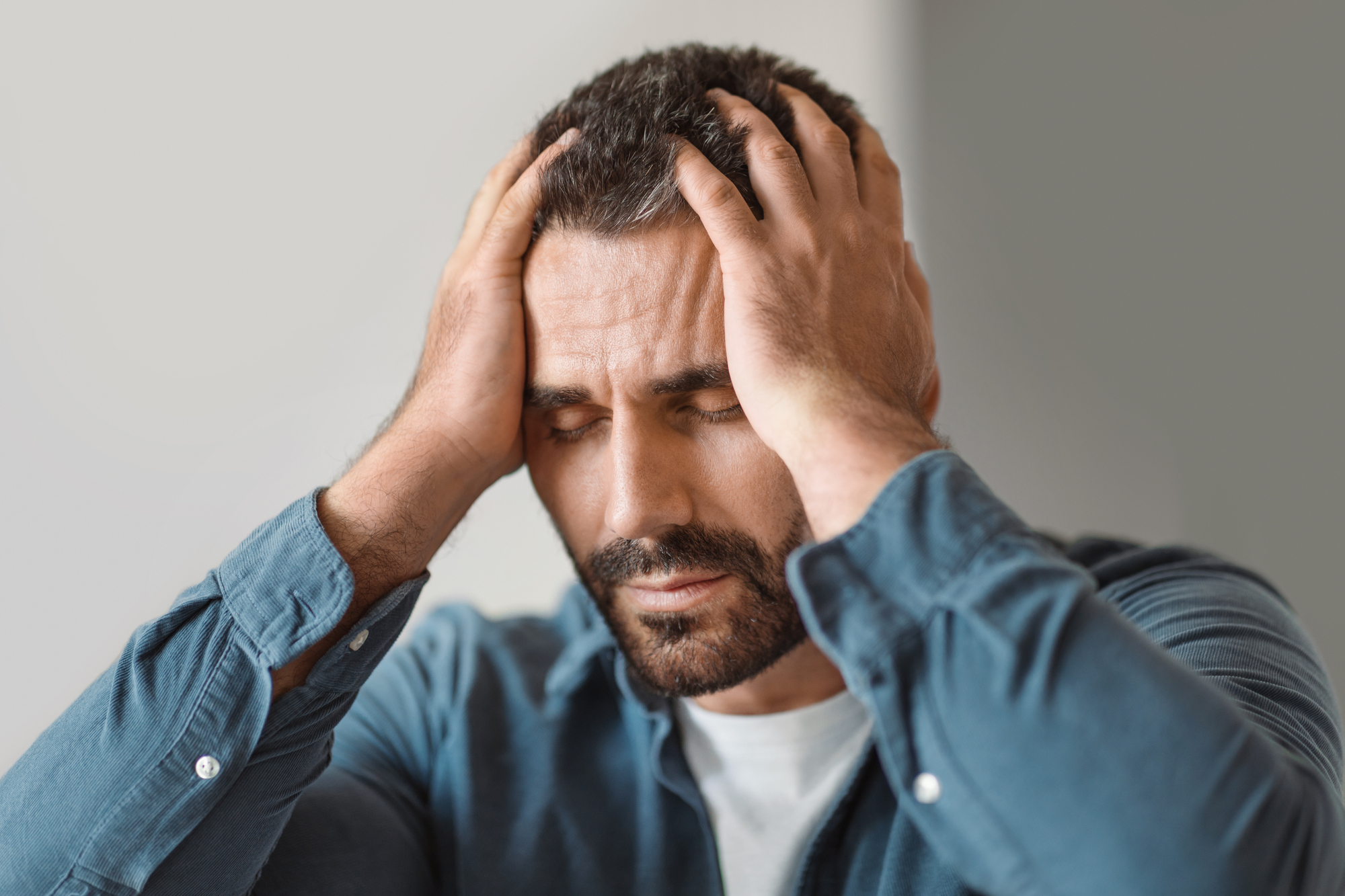 A man with a beard and closed eyes holds his head in his hands, appearing stressed or in pain. He is wearing a blue shirt and is positioned in front of a neutral background.