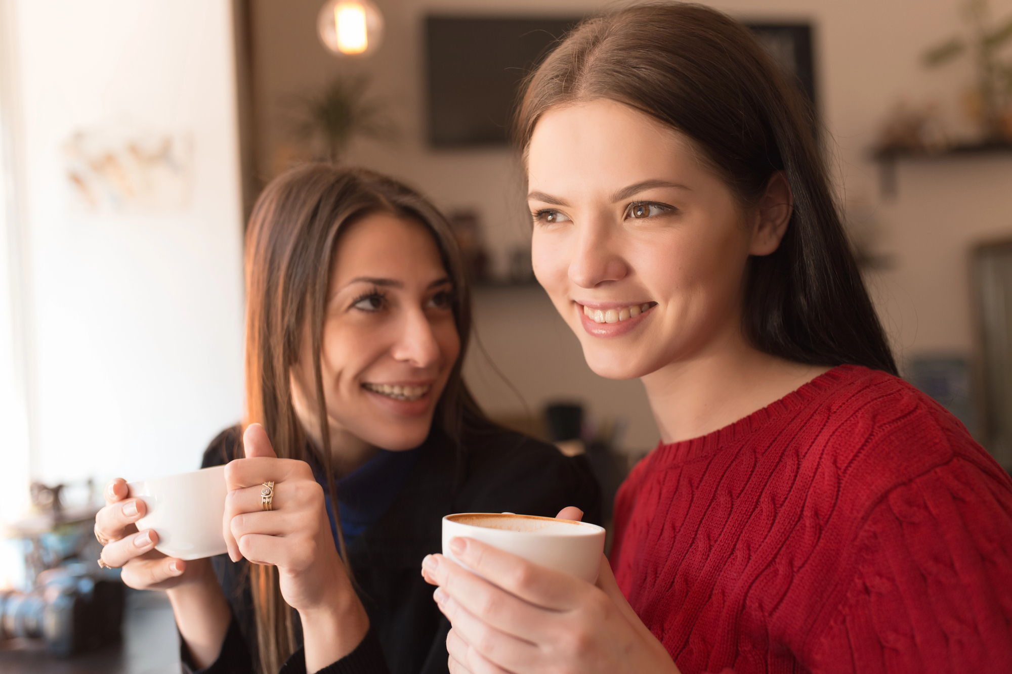 Two women are sitting together in a cozy cafe, holding cups of coffee. One woman is wearing a red sweater and smiling, while the other is looking at her with a smile. The background is softly blurred, creating a warm atmosphere.